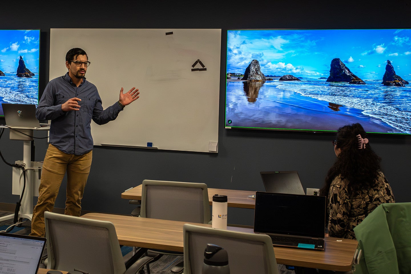 Masc presenting student with glasses and a goatee in a button down, long sleeve shirt presents to a crowded room during midterms in the UO library presentation class room. 